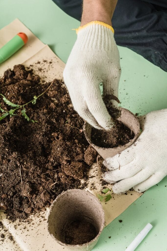 Close-up of a person's hands wearing white gardening gloves, carefully filling a small, round, sustainable paper pot with rich, dark soil on a green workspace, with gardening tools and a scattering of soil visible around